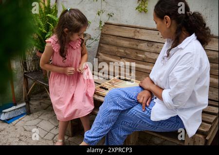 Mutter und Tochter teilen einen fröhlichen Moment, als sie draußen auf einer Holzbank Backgammon spielen. Die Szene erinnert an Familie, Bindung und Freizeit. Stockfoto