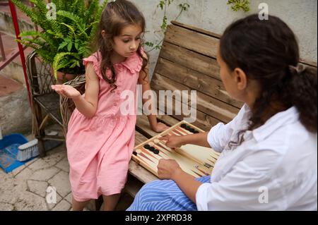 Mutter und Tochter genießen gute Zeit beim Spielen von Backgammon im Freien auf einer rustikalen Holzbank. Das Kind in einem rosa Kleid hört aufmerksam zu. Stockfoto