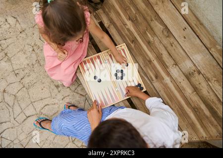 Mutter und Tochter spielen zusammen ein Spiel Backgammon draußen. Die Holzbank und der Steinboden verstärken die gemütliche und entspannende Atmosphäre. Stockfoto