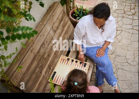 Blick von oben auf eine Mutter und Tochter, die Backgammon auf einer Holzbank in einem Garten spielen. Familienbeziehungen und Freizeitaktivitäten. Stockfoto