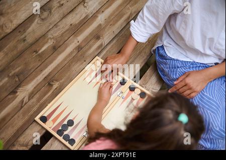 Blick von oben auf Eltern und Kinder, die gemeinsam im Freien ein Spiel Backgammon spielen. Sie sitzen auf einer Holzbank und genießen eine gute Familienzeit. Stockfoto