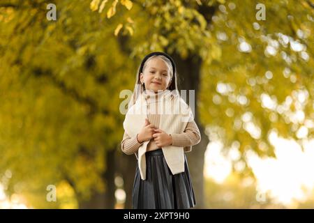 Kindertag. Fröhliches kleines Mädchen 5-6 Jahre alt posiert und lächelt draußen vor der Kamera und steht am Baum im Herbstpark. Das stilvolle, zarte Vorschulkind trägt einen beigefarbenen Strickpullover. Glückliche Kindheit Stockfoto