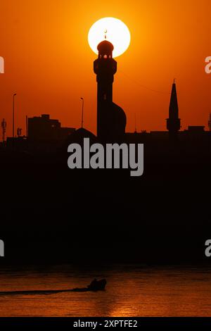 Ein Boot, das während des Sonnenuntergangs auf dem Tigris hinter dem Minarett der Imam Mohsen Moschee in der Stadt Mossul im Nordirak segelt. Stockfoto