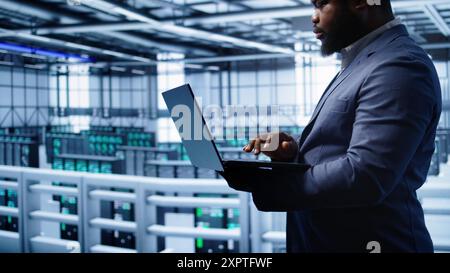 Softwareentwickler im Server-Hub prüfen den Wiederherstellungsplan auf dem Laptop und überwachen den Energieverbrauch der Hardware. afroamerikanischer Mitarbeiter verwendet Notebook im Rechenzentrum, um sicherzustellen, dass die Rigs funktionieren, Kamera B Stockfoto