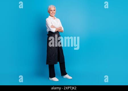 Ganzkörperfoto einer attraktiven Seniorin überkreuzte Hände kleine Business Café Besitzer gekleidete Kellnerin Uniform isoliert auf blauem Hintergrund Stockfoto