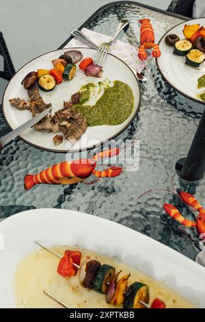 Essen von Steak-Scheiben mit grüner Chimichurri-Sauce und gegrilltem Gemüse auf Tellern am Esstisch im Freien Stockfoto
