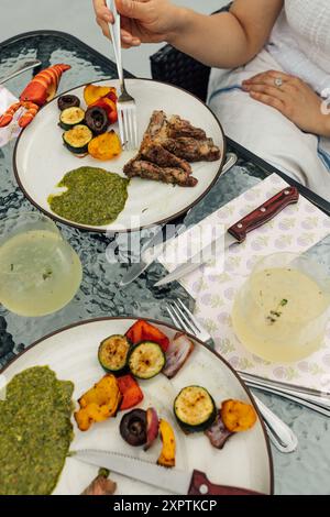 Essen von Steak-Scheiben mit grüner Chimichurri-Sauce und gegrilltem Gemüse auf Tellern am Esstisch im Freien Stockfoto