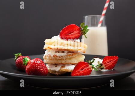 Ein Stapel Pfannkuchen mit Sahne und Erdbeeren auf einem schwarzen Teller, mit einem Glas Milchshake im Hintergrund Stockfoto