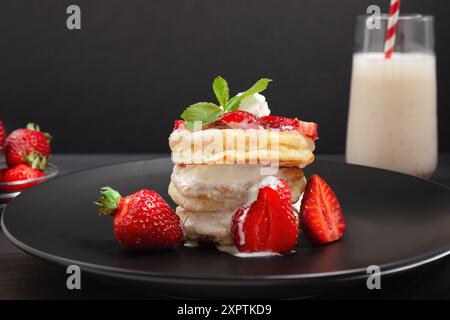 Ein Stapel flauschiger Pfannkuchen mit frischen Erdbeeren und Schlagsahne, serviert mit einem Glas Milchshake auf einem schwarzen Teller. Stockfoto