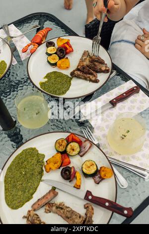 Essen von Steak-Scheiben mit grüner Chimichurri-Sauce und gegrilltem Gemüse auf Tellern am Esstisch im Freien Stockfoto