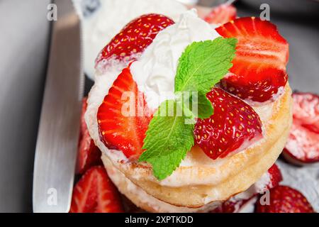 Detaillierte Ansicht von flauschigen Pfannkuchen mit frischen Erdbeeren, Schlagsahne und Minzblättern, präsentiert auf einem Teller mit einem Messer, unordentliches Essen Stockfoto