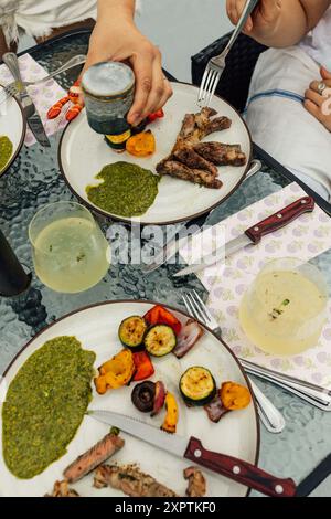 Essen von Steak-Scheiben mit grüner Chimichurri-Sauce und gegrilltem Gemüse auf Tellern am Esstisch im Freien Stockfoto