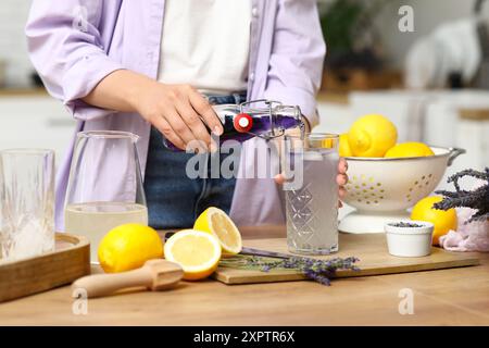 Junge Frau, die Lavendelsaft aus der Flasche in das Glas gießt, um Limonade am Tisch in der Küche zuzubereiten Stockfoto