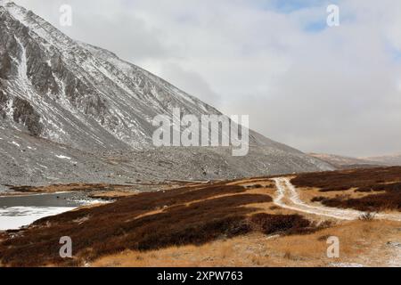 Eine Feldstraße, die mit dem ersten Schnee bedeckt ist, überquert an einem bewölkten Herbsttag einen mit gelblichem Gras bewachsenen Hügel am Fuße einer hohen Klippe. Naryn-Gol ri Stockfoto