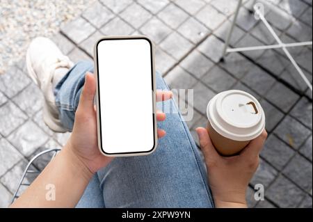 Draufsicht einer Frau in Jeans, die im Freien sitzt und mit einer Kaffeetasse in der Hand ihr Smartphone benutzt und in einem Café kühlt. Ein Smartphone wi Stockfoto
