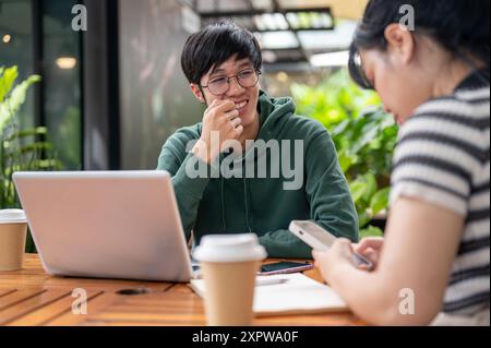 Ein junger, glücklicher asiatischer Student in einem grünen Hoodie macht Spaß mit seinem Freund zu reden, während er gemeinsam in einem Café Hausaufgaben macht. Stockfoto