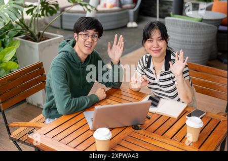 Zwei fröhliche, freundliche junge asiatische Freundinnen sitzen draußen an einem Tisch und winken mit der Hand zur Kamera. Freundschaft, Teamarbeit, Stockfoto