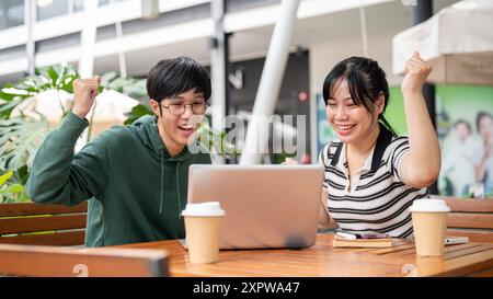 Zwei fröhliche und aufgeregte junge asiatische Freunde, männlich und weiblich, schauen auf einen Laptop-Bildschirm, überrascht von den guten Nachrichten und feiern zusammen, was sie feiern Stockfoto