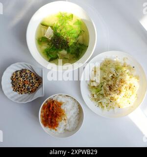 Vietnamesische Mahlzeit mit gedämpftem Reis, Tofu und Gemüsesuppe, gebratenen grünen Bohnen, Bambussprossen und Chili-Sauce auf einem weißen Tisch, beleuchtet von na Stockfoto