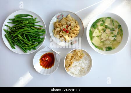 Vietnamesische Mahlzeit mit gedämpftem Reis, Tofu und Gemüsesuppe, gebratenen grünen Bohnen, Bambussprossen und Chili-Sauce auf einem weißen Tisch, beleuchtet von na Stockfoto