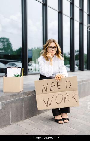 Eine Frau, die draußen mit einem Pappbanner mit dem Slogan sitzt, braucht Arbeit neben einer Schachtel. Schöne weibliche Demonstrantin im Kampf gegen die Arbeitslosigkeit. Stockfoto