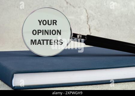 Textschild, das IHRE MEINUNG durch eine Lupe auf einem Business-Notebook vor einem Wandhintergrund anzeigt Stockfoto