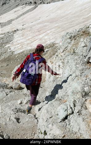 Junge Frau mit violettem Rucksack, die im Sommer auf einem steilen, felsigen Bergweg unterwegs ist Stockfoto