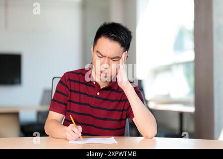 Ein junger männlicher Student im Teenageralter macht eine Prüfung oder Hausaufgaben in einem Klassenzimmer und sieht gestresst aus, während er mit einem Bleistift auf den Schreibtisch schreibt Stockfoto