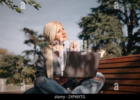 Eine blonde Frau sitzt auf einer Bank mit einem Laptop vor sich. Sie trägt eine graue Jacke und Jeans. Die Szene deutet auf eine lässige und entspannte Atmosphäre hin Stockfoto