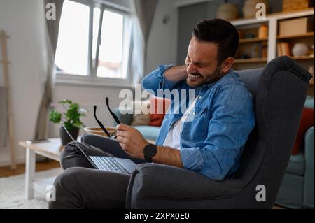 Der gestresste Webdesigner leidet unter Nackenbart, der eine Brille hält und mit einem Laptop auf dem Sessel sitzt. Junger Unternehmer, der seine Augen vor Schmerzen schließt Stockfoto