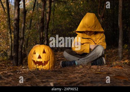 Ein nicht erkennbares Kind mit einer orangefarbenen Kapuze über dem Gesicht sitzt auf dem Boden, bestreut mit gefallenen Blättern neben einem großen geschnitzten Halloween-Kürbis. Stimmung Stockfoto