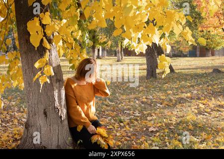 Ein nicht erkennbares erwachsenes Mädchen in einem gelben Pullover sitzt auf dem Boden, bedeckt mit leuchtenden, herbstlich gelb-orangen Blättern, ihr Gesicht ist mit Haaren bedeckt. Sonnige Farbe Stockfoto