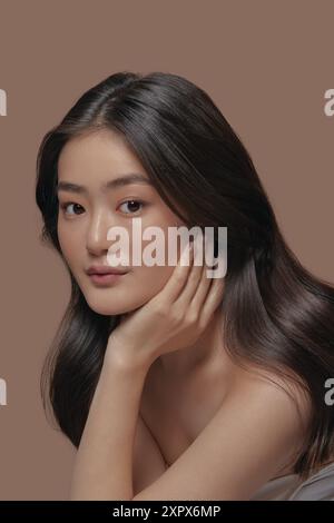 Charmante junge asiatische Frau mit einer gesunden, sauberen Haut neigt sich die Hand zum Gesicht und posiert über beigefarbenen isolierten Hintergrund, vertikales Nahfoto. Stockfoto