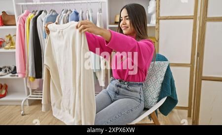 Eine junge hispanische Frau untersucht einen weißen Pullover in einem hell beleuchteten Ankleidezimmer und zeigt alltägliche Mode. Stockfoto