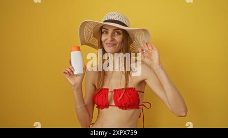 Junge, attraktive Frau in rotem Bikini und Strohhut mit Sonnencreme, lächelnd über einem isolierten gelben Hintergrund. Stockfoto