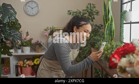 Lächelnde hispanische Frau fotografiert Pflanzen mit einem Telefon in einem Blumenladen. Stockfoto