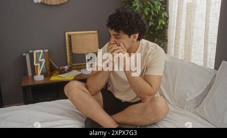 Ein junger, attraktiver hispanischer Mann sitzt nachdenklich auf seinem Bett in einem gemütlichen, modernen Schlafzimmer, das von natürlichem Licht durch das Fenster beleuchtet wird. Stockfoto