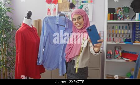 Lächelnde Frau im Hijab, die Selfie im Schneiderladen mit Schaufensterpuppe und bunten Fäden im Hintergrund macht. Stockfoto