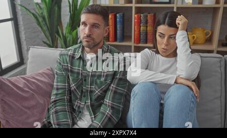 Ein hispanischer Mann und eine hispanische Frau zeigen sich besorgt, während sie zusammen auf einem grauen Sofa in einem gut eingerichteten Wohnzimmer sitzen. Stockfoto