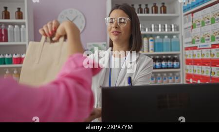 Eine Apothekerin in einem weißen Mantel hilft einem Kunden in Rosa an einer Apotheke mit verschiedenen Medikamenten und Produkten in den Regalen im Hintergrund Stockfoto