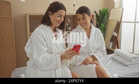 Zwei hispanische Frauen in weißen Bademänteln sitzen zusammen in einem Spa und genießen ihre Zeit, während sie auf ein Telefon schauen. Stockfoto