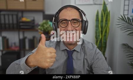 Ein hispanischer Mann mittleren Alters in einem Büro, der die Daumen hochlegt, während er Kopfhörer und Brille trägt. Stockfoto