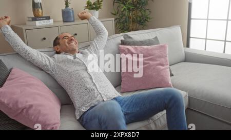 Hispanischer Mann mittleren Alters, der sich auf einer grauen Couch in einem modernen Wohnzimmer entspannt und entspannt. Stockfoto