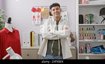 Selbstbewusster männlicher Modedesigner mit überkreuzten Armen in einem Schneidergeschäft, umgeben von Schaufensterpuppen, Stoff und Nähgarnen. Stockfoto