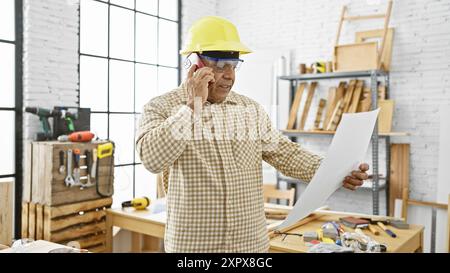 Reifer hispanischer Mann mit Helm mit Telefon in einer gut ausgestatteten Tischlerei. Stockfoto