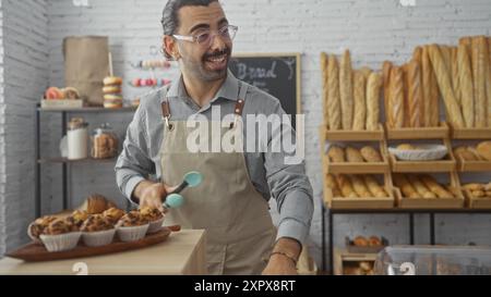 Junger Mann mit Schnurrbart, der in einer Bäckerei mit frischem Brot und Gebäck arbeitet und lächelt, während er Muffins von der Theke mit einer Zange serviert Stockfoto