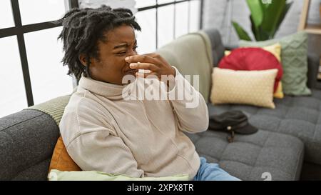 Eine junge afroamerikanerin mit Dreadlocks in einem Rollkragen, sitzt drinnen auf einem Sofa und kneift ihre Nase vor Unbehagen. Stockfoto