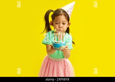 Fokussiertes fünfjähriges chinesisches Aussehen Kind in Party Hut Blow Out Kerzen auf Geburtstagskuchen hält es und steht vor gelbem Hintergrund. Stockfoto