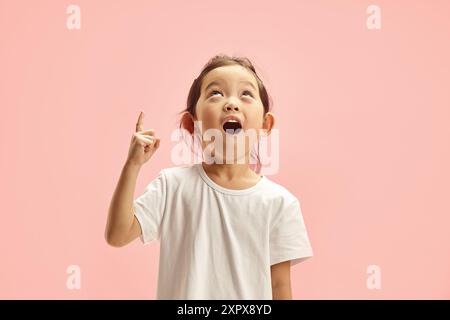 Positive asiatische kleine Mädchen Punkte oben zeigen für Ihre Werbetext Lächeln überrascht trägt weißes T-Shirt isoliert über rosa Hintergrund. Stockfoto