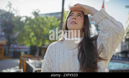 Eine junge hispanische Frau in einem weißen Pullover schaut nach oben, während sie ihre Augen draußen in einem sonnigen Park abschirmt. Stockfoto